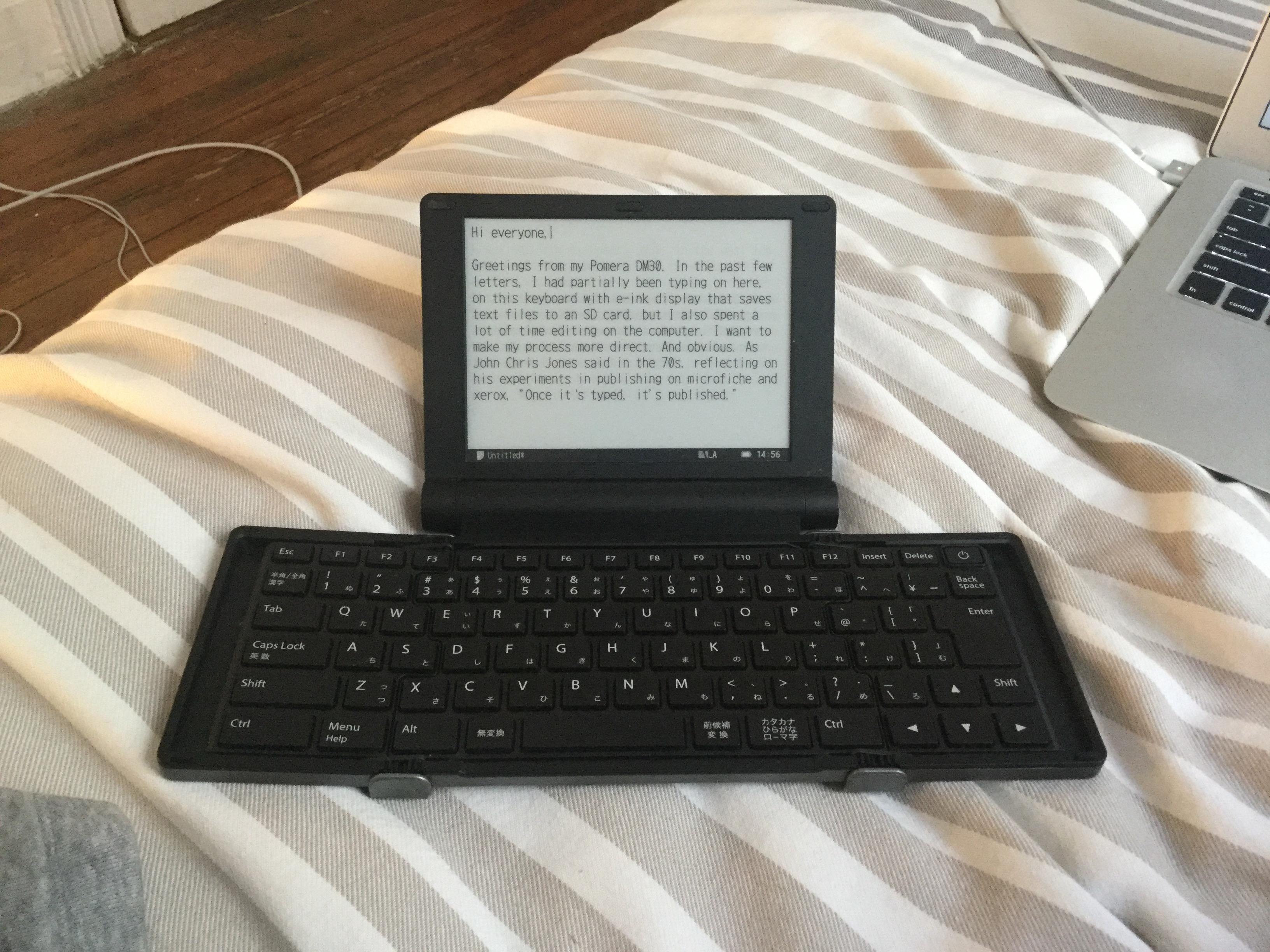 Photograph of a Pomera DM30, a small computer-like device made in Japan that has an e-ink display that is intended to be a distraction-free writing environment, on a bed. The corner of a laptop is seen in the upper right.