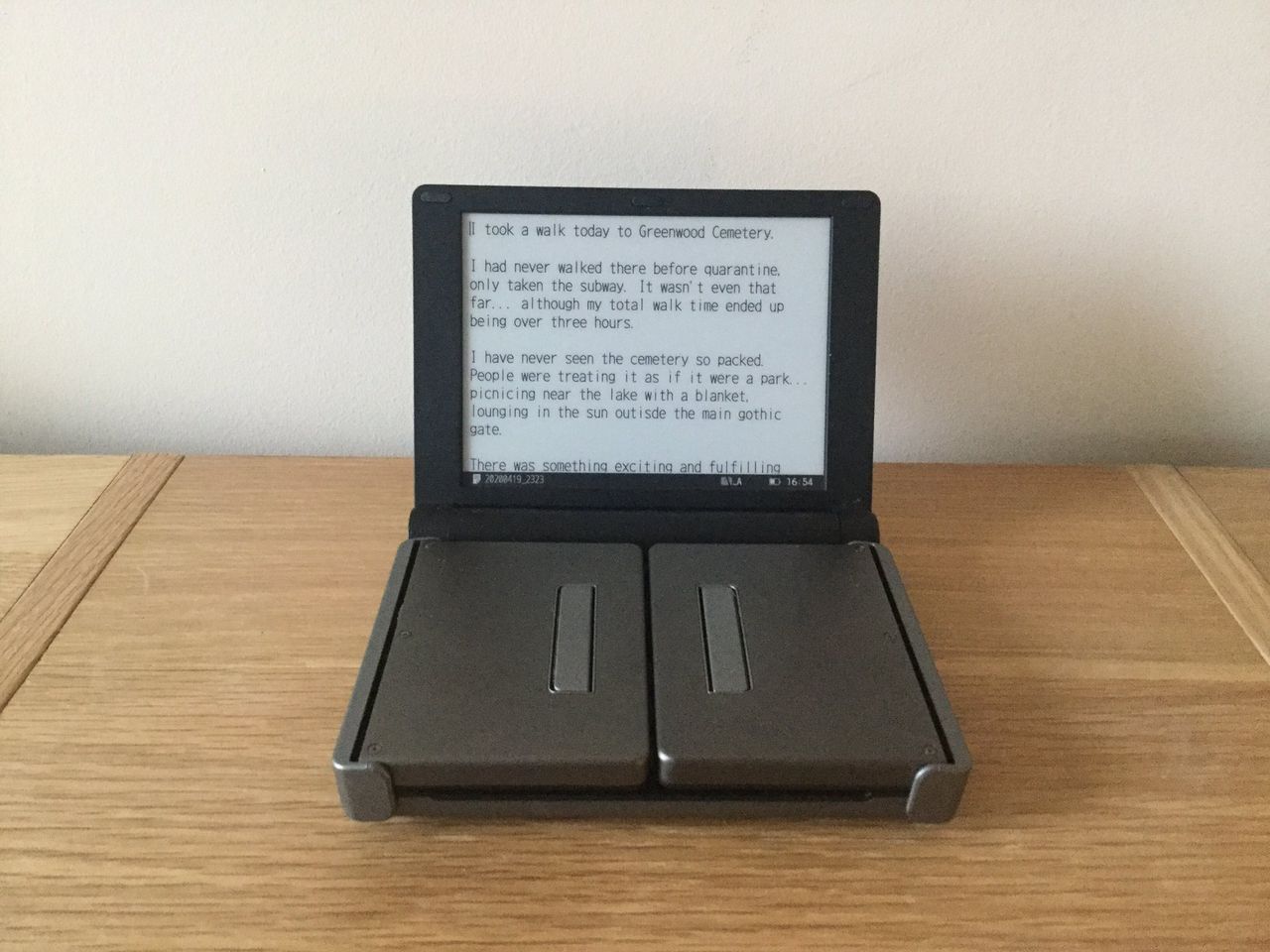 The Pomera DM30 on a wooden surface, partially unfolded but hiding its keyboard. It has text on it, starting with 'I took a walk today to Greenwood Cemetery...'